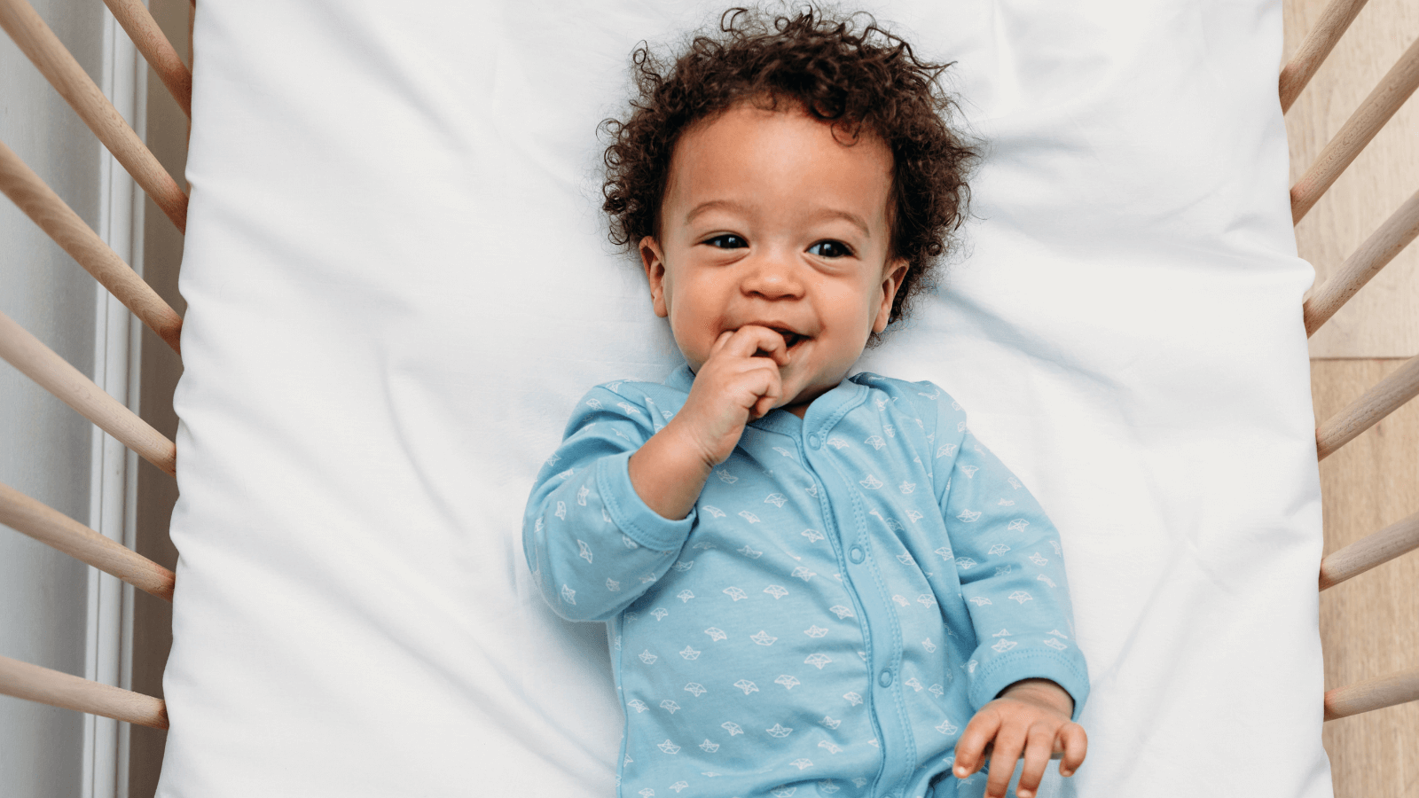 Happy baby with curly hair wearing a blue onesie lying in a crib, smiling and sucking on fingers.