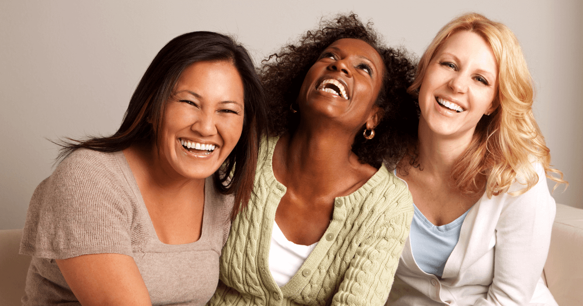 Three diverse women sitting together on a couch, laughing and smiling.