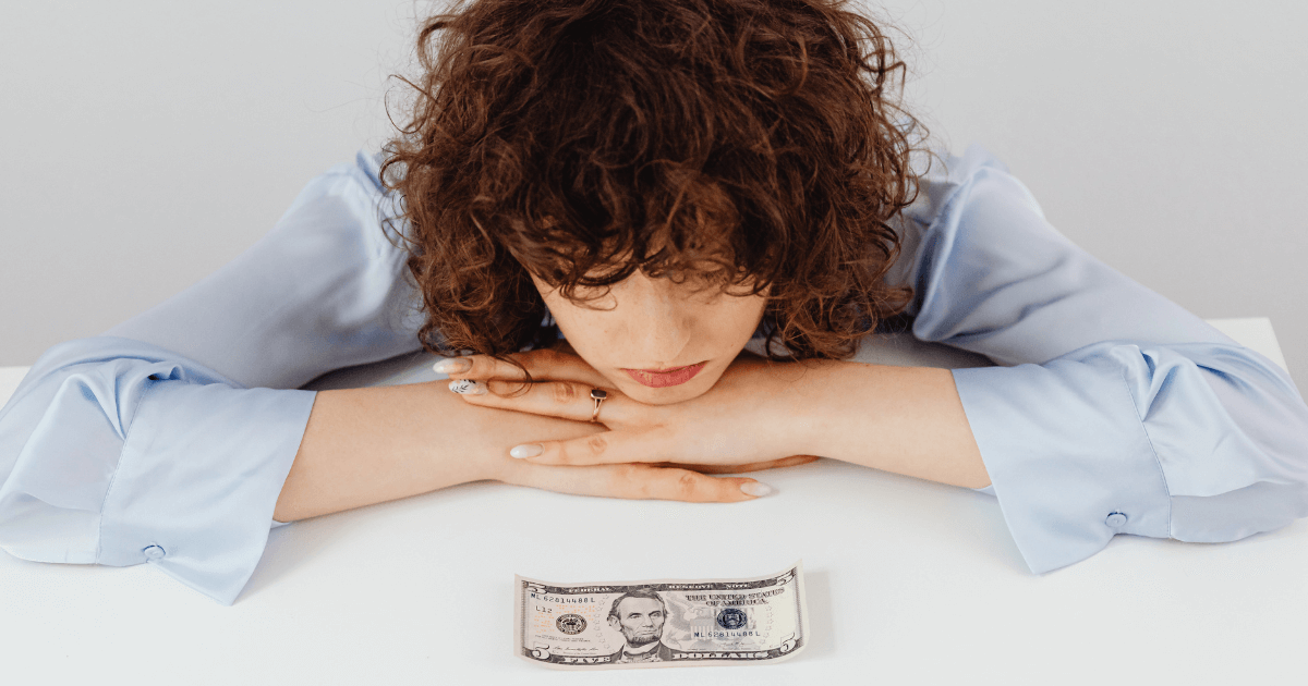 Why Some Nannies Work Off the Books (And Why Some Don’t) - Young woman looking down at a five-dollar bill on a white table, appearing worried.