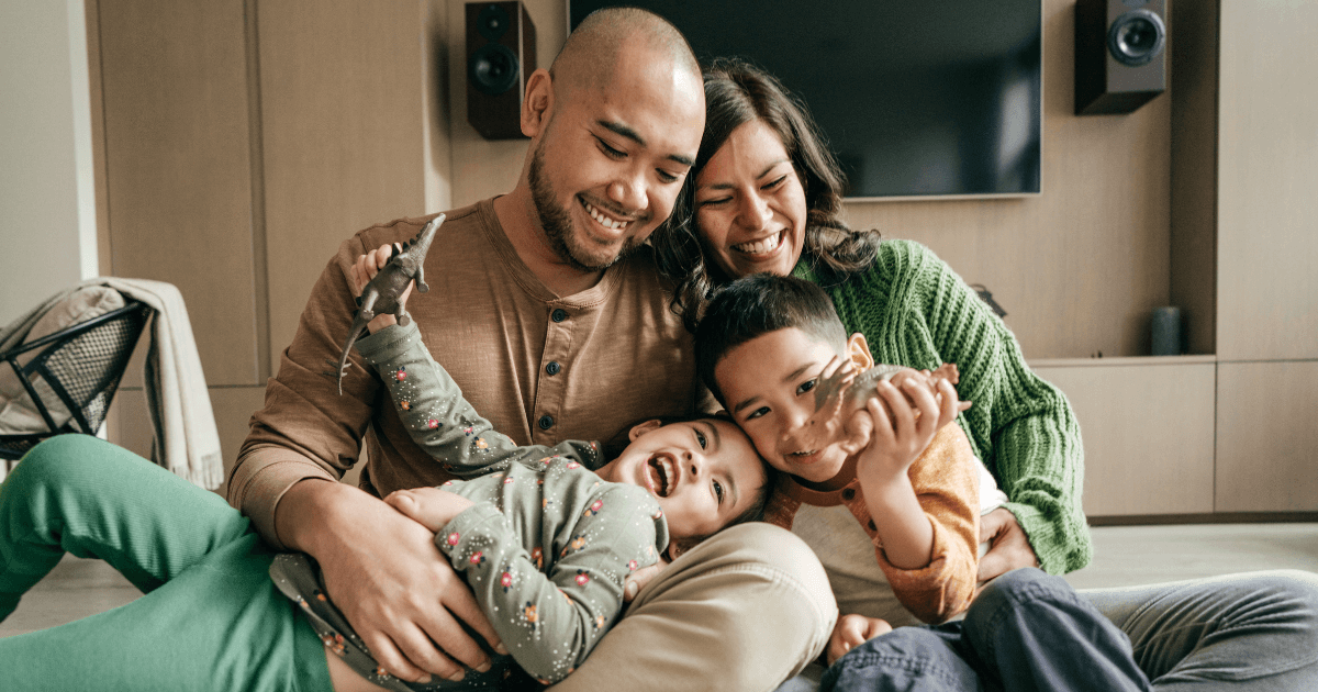 Smiling parents hugging two children while playing with toy dinosaurs at home.